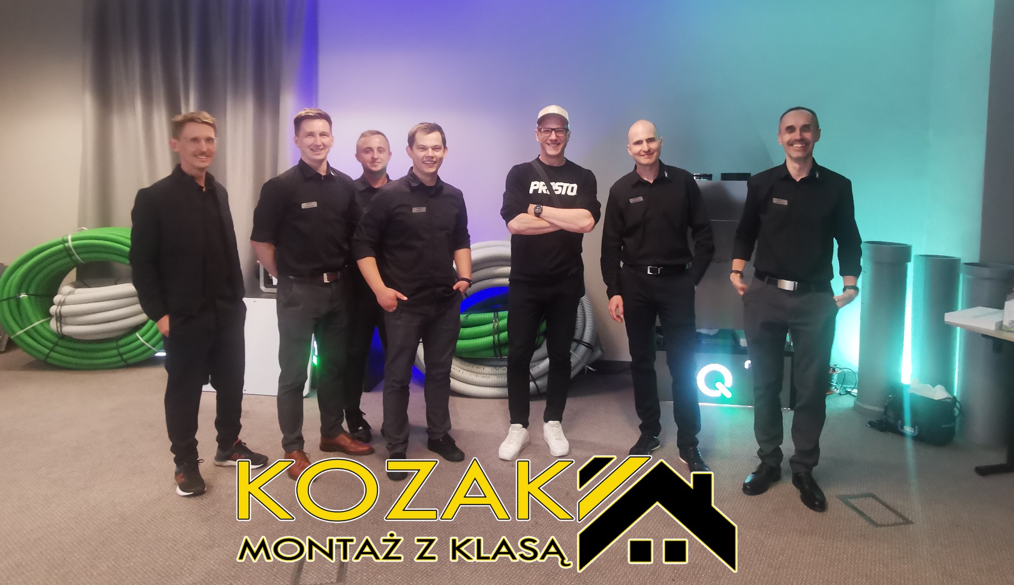Zespół monterów w czarnych uniformach pozuje na tle zwiniętych rur wentylacyjnych i szarych rur kanalizacyjnych, widoczne logo firmy 'KOZAK Montaż z Klasą'.