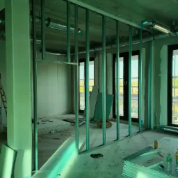 Montaż metalowej konstrukcji ścian działowych w surowym stanie, widoczne płyty gipsowo-kartonowe, instalacja wentylacyjna pod sufitem, okna z widokiem na zieleń.