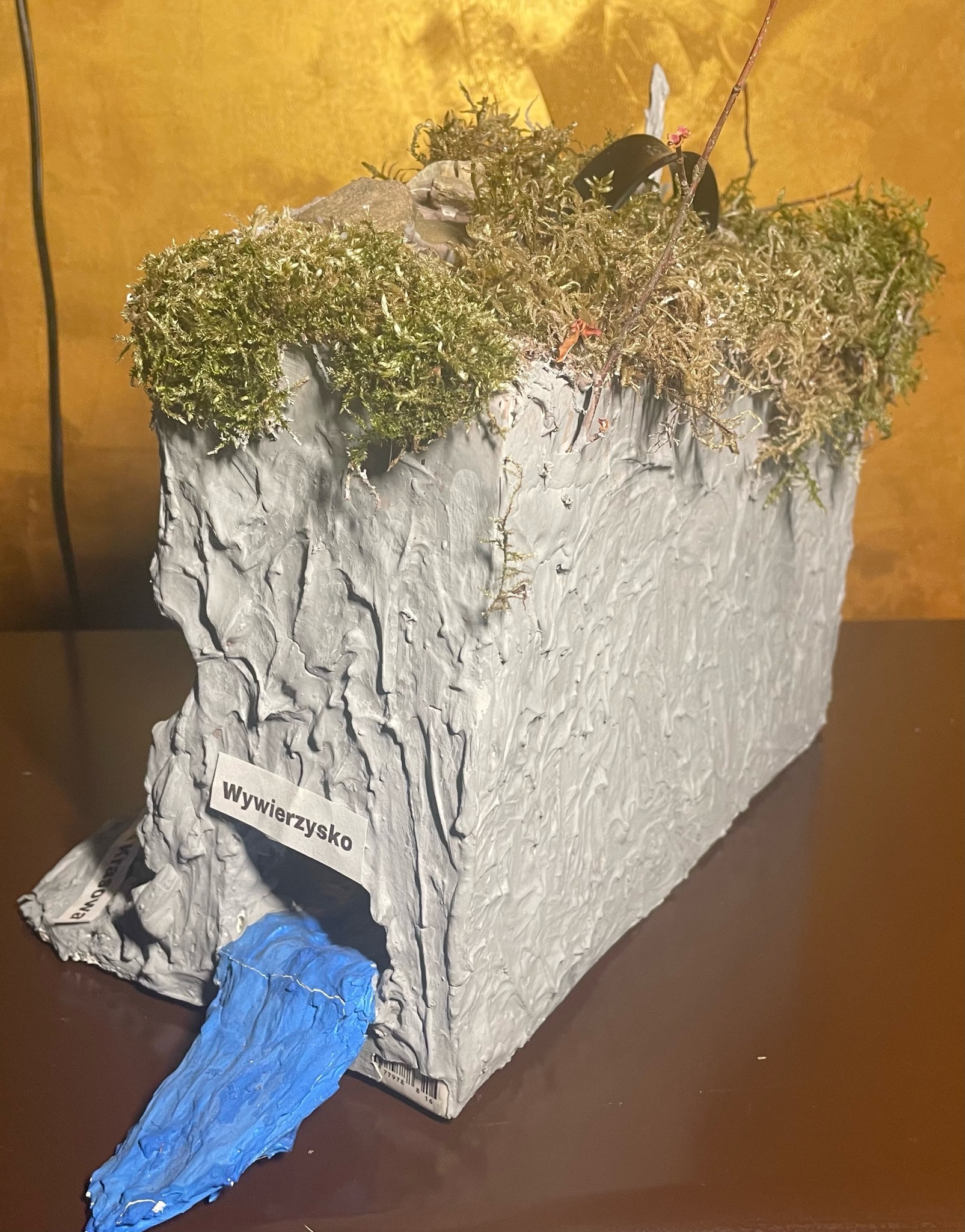 Makieta groty z szarego materiału z mchem na górze, imitacją wodospadu z niebieskiego materiału i napisem 'Wywierżysko'