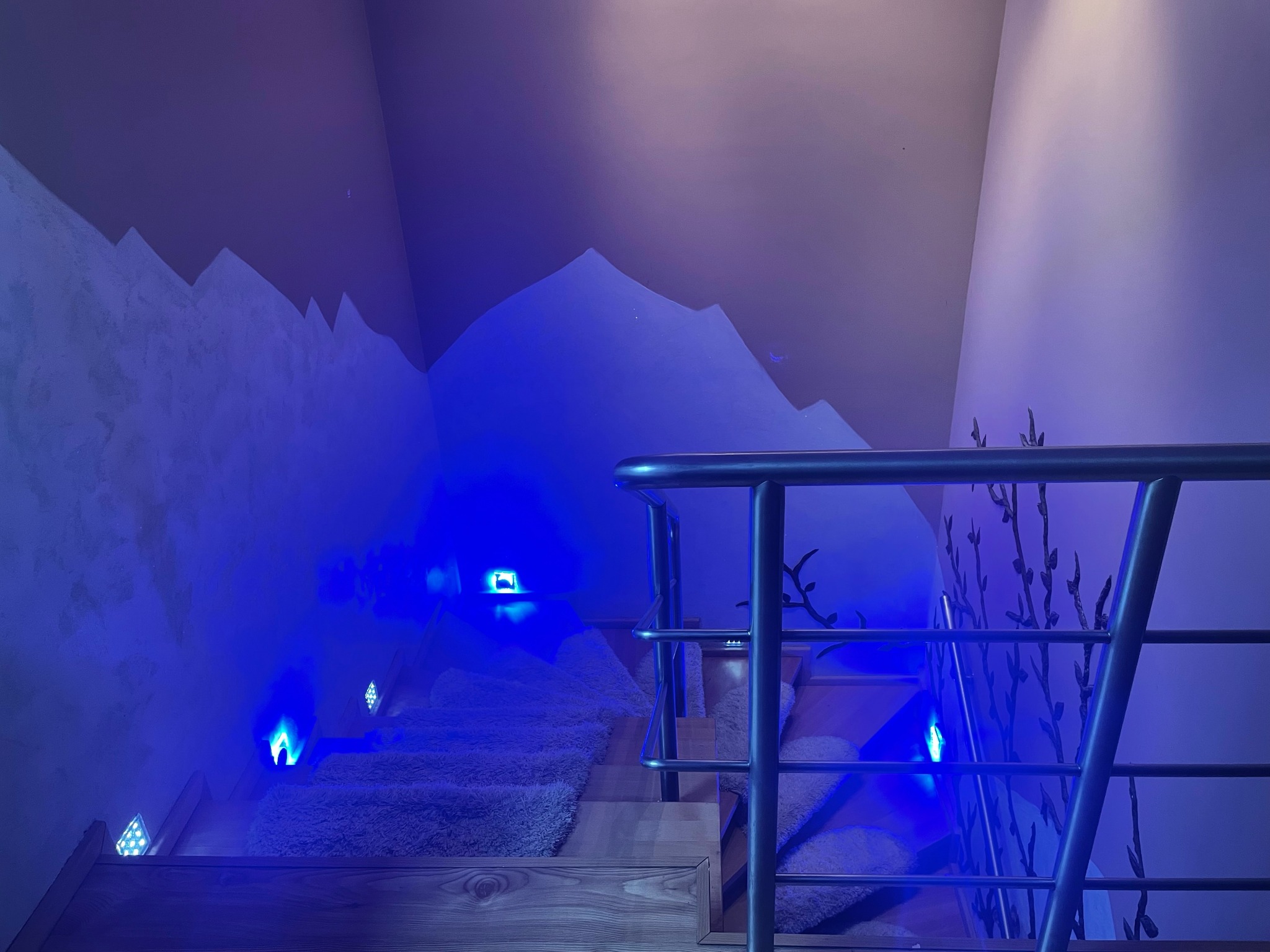 Schody wewnętrzne z drewnianymi stopniami pokryte puszystym dywanem, oświetlone niebieskimi lampkami LED, z dekoracją ścienną przedstawiającą górski krajobraz, widziane z dołu przez barierki.