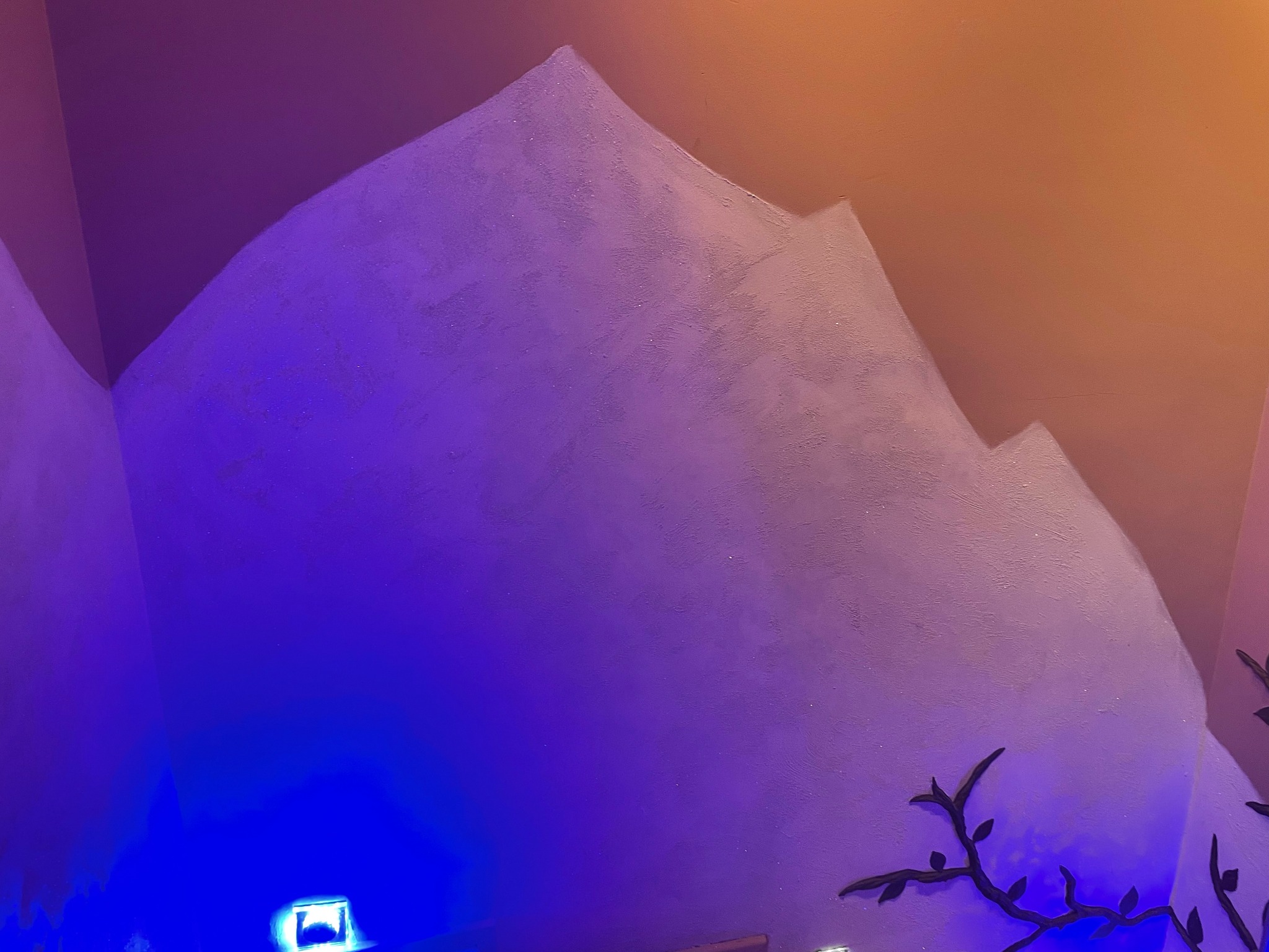 Ściana z ręcznie malowanym motywem górskim w odcieniach fioletu i beżu, oświetlona niebieskim światłem punktowym i zdobiona ciemnymi gałązkami.