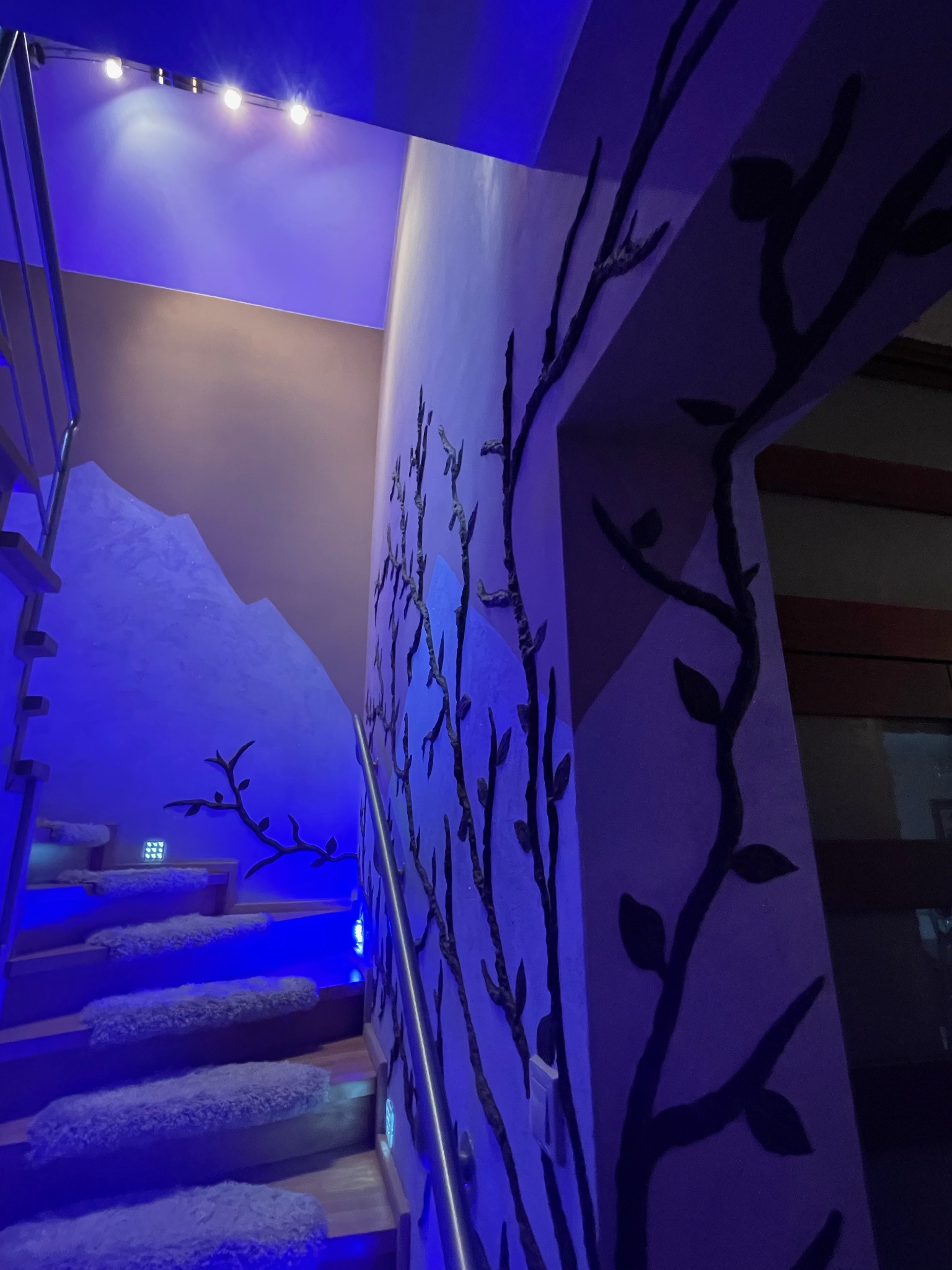 Schody z drewnianymi stopniami pokrytymi białym futerkiem, oświetlone niebieskim światłem LED, ściana ozdobiona czarnymi, reliefowymi gałęziami z liśćmi, w tle imitacja górskiego krajobrazu.