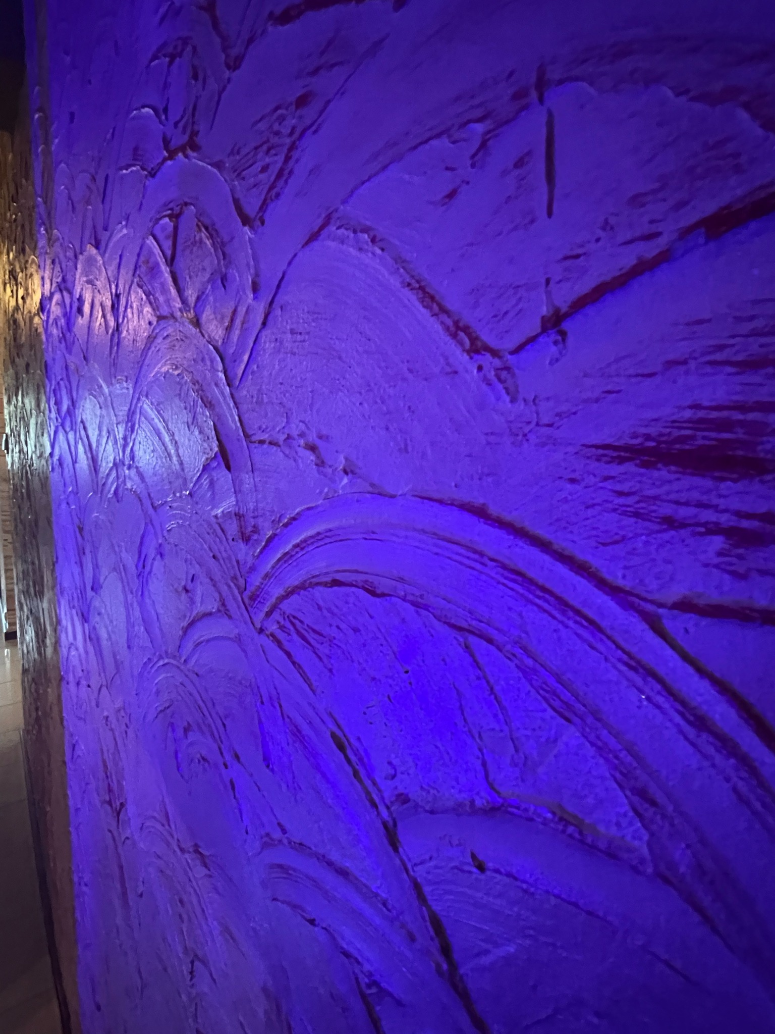 Teksturowana ściana pomalowana na fioletowo, z widocznymi śladami pędzla i nierównościami, oświetlona intensywnym fioletowym światłem.