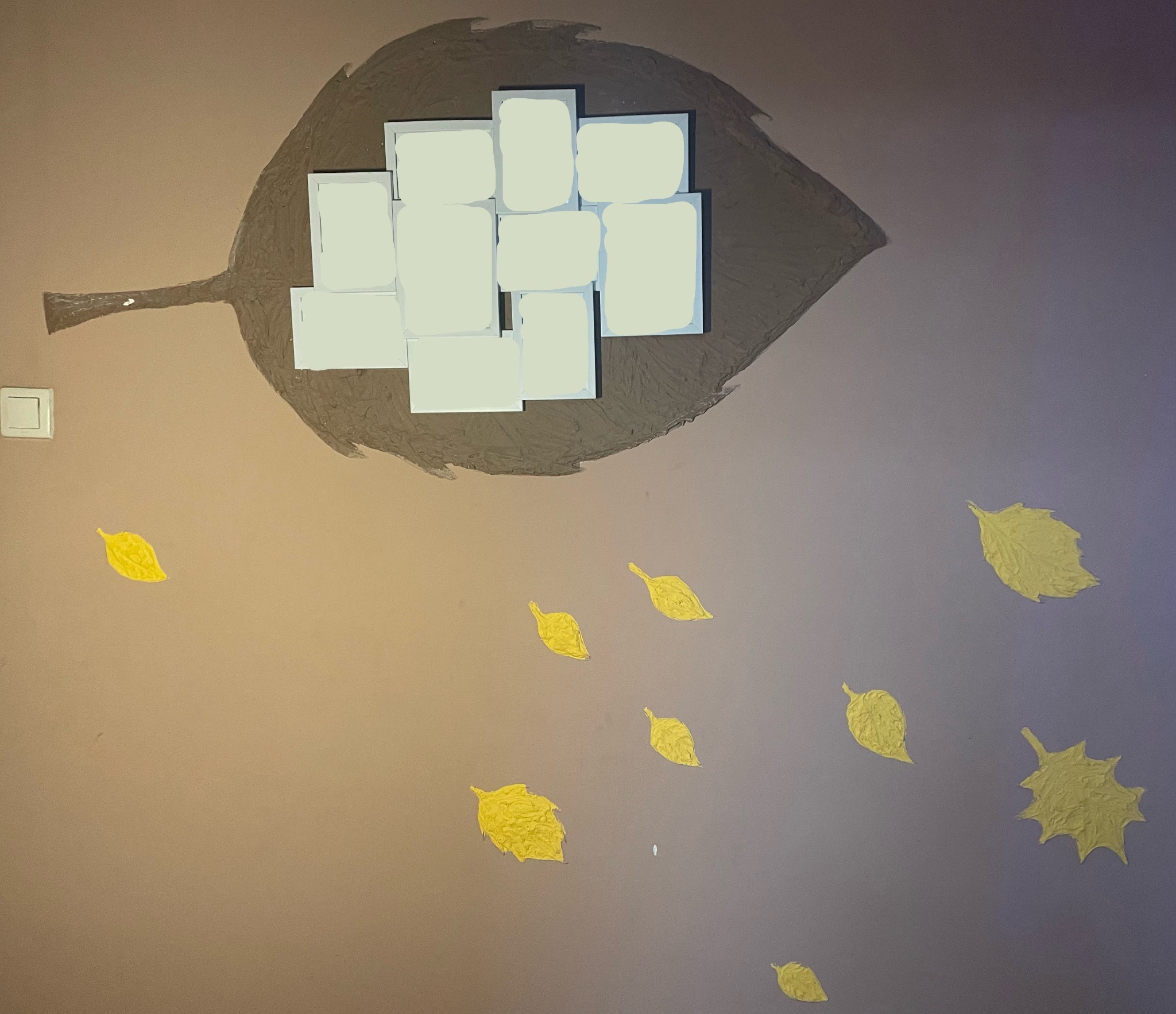 Brązowy motyw liścia z białymi kwadratami wewnątrz na beżowej ścianie, ozdobiony złotymi liśćmi opadającymi w dół, obok włącznika światła.