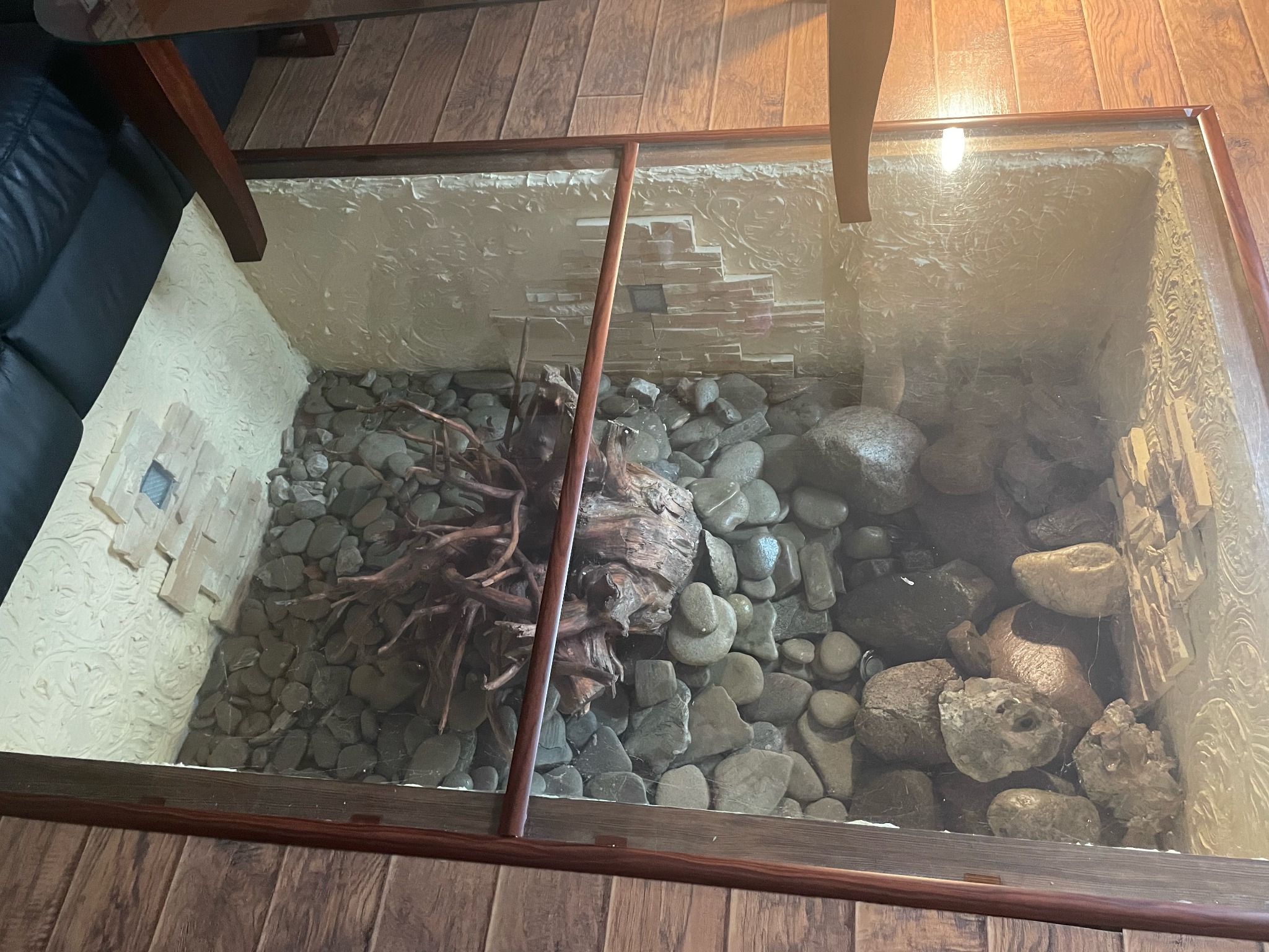 Stolik kawowy ze szklanym blatem, pod którym znajduje się kompozycja z kamieni, korzeni i dekoracyjnej ściany z efektem murowanej cegły.