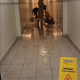 EVERY ON SPÓŁKA Z OGRANICZONĄ ODPOWIEDZIALNOŚCIĄ - Pracownicy w uniformach czyszczą mokrą podłogę w korytarzu za pomocą maszyny polerującej; żółty znak ostrzegawczy o śliskiej podłodze na pierwszym planie.