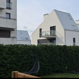 EVERY ON SPÓŁKA Z OGRANICZONĄ ODPOWIEDZIALNOŚCIĄ - Nowoczesne, białe budynki mieszkalne z szarymi dachami, częściowo zasłonięte przez wysoki, zielony żywopłot.