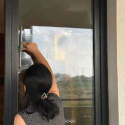 EVERY ON SPÓŁKA Z OGRANICZONĄ ODPOWIEDZIALNOŚCIĄ - Kobieta z upiętymi włosami, używa ściągaczki do mycia dużej, panoramicznej szyby okiennej z ciemną ramą, widok z wnętrza na zewnątrz.