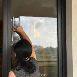 Kobieta z upiętymi włosami, używa ściągaczki do mycia dużej, panoramicznej szyby okiennej z ciemną ramą, widok z wnętrza na zewnątrz.