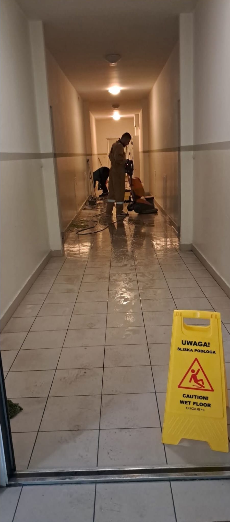 Pracownicy w uniformach czyszczą mokrą podłogę w korytarzu za pomocą maszyny polerującej; żółty znak ostrzegawczy o śliskiej podłodze na pierwszym planie.