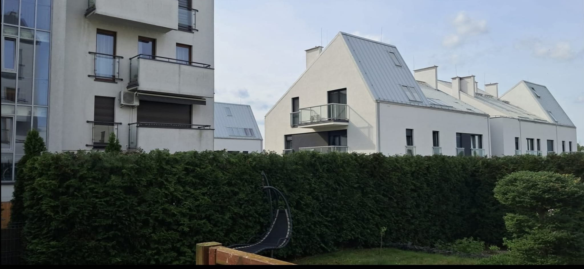 Nowoczesne, białe budynki mieszkalne z szarymi dachami, częściowo zasłonięte przez wysoki, zielony żywopłot.