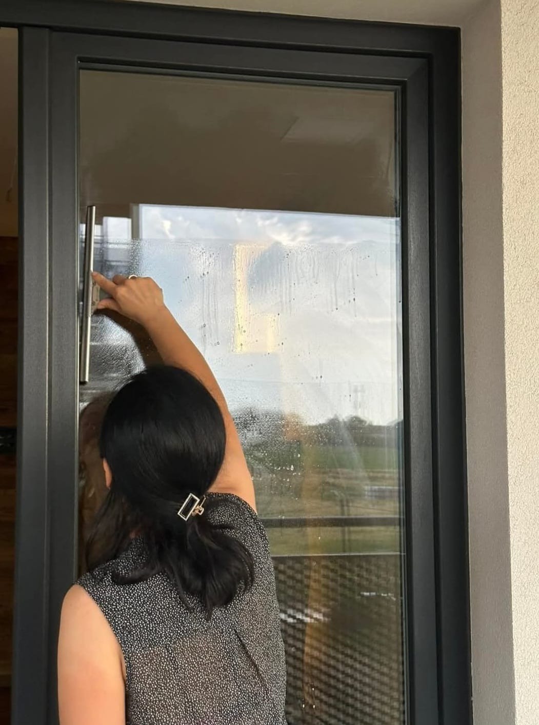 Kobieta z upiętymi włosami, używa ściągaczki do mycia dużej, panoramicznej szyby okiennej z ciemną ramą, widok z wnętrza na zewnątrz.