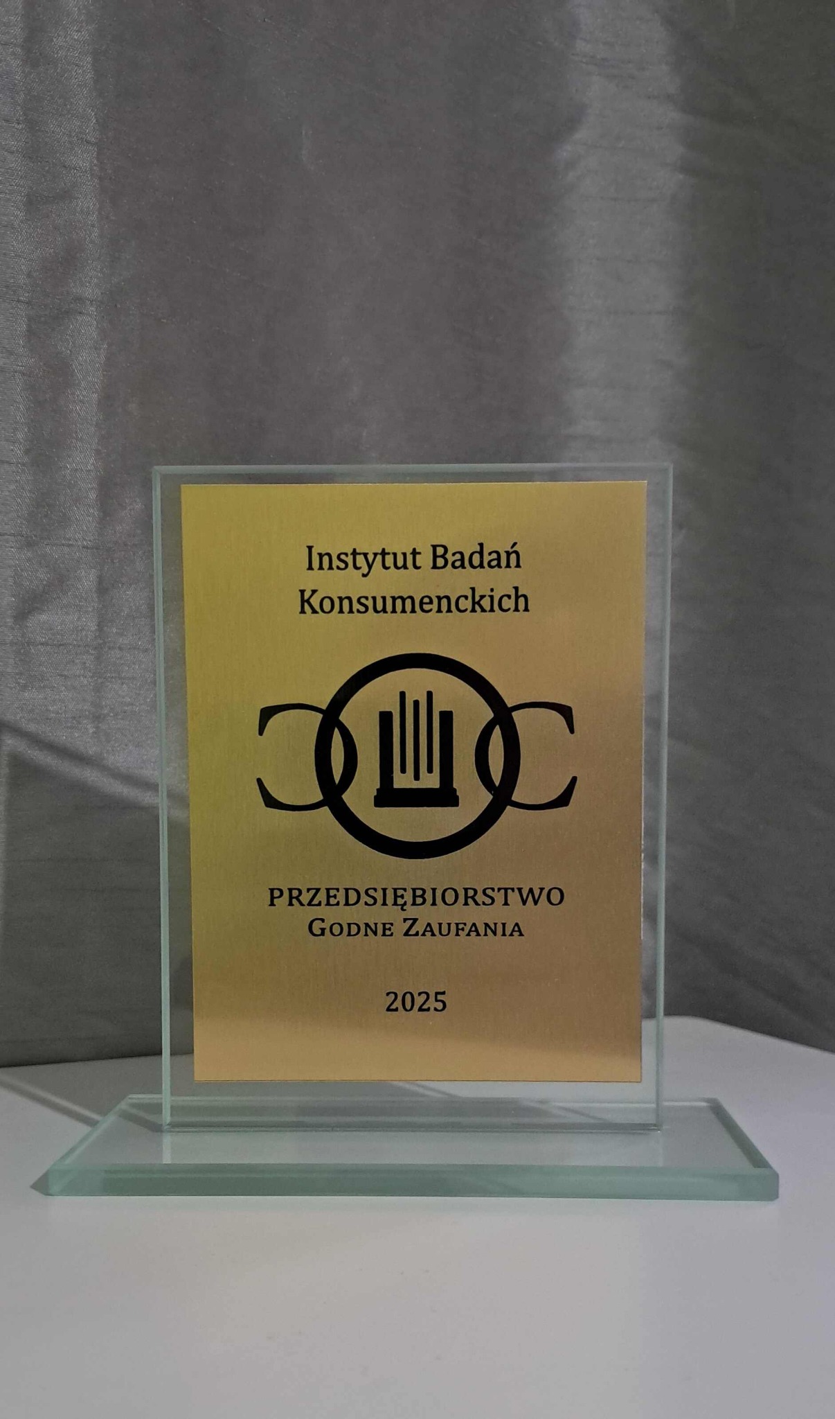 Szklana statuetka z napisem 'Instytut Badań Konsumenckich Przedsiębiorstwo Godne Zaufania 2025' na złotym tle. Prestiżowa nagroda na białym blacie, szare tło.