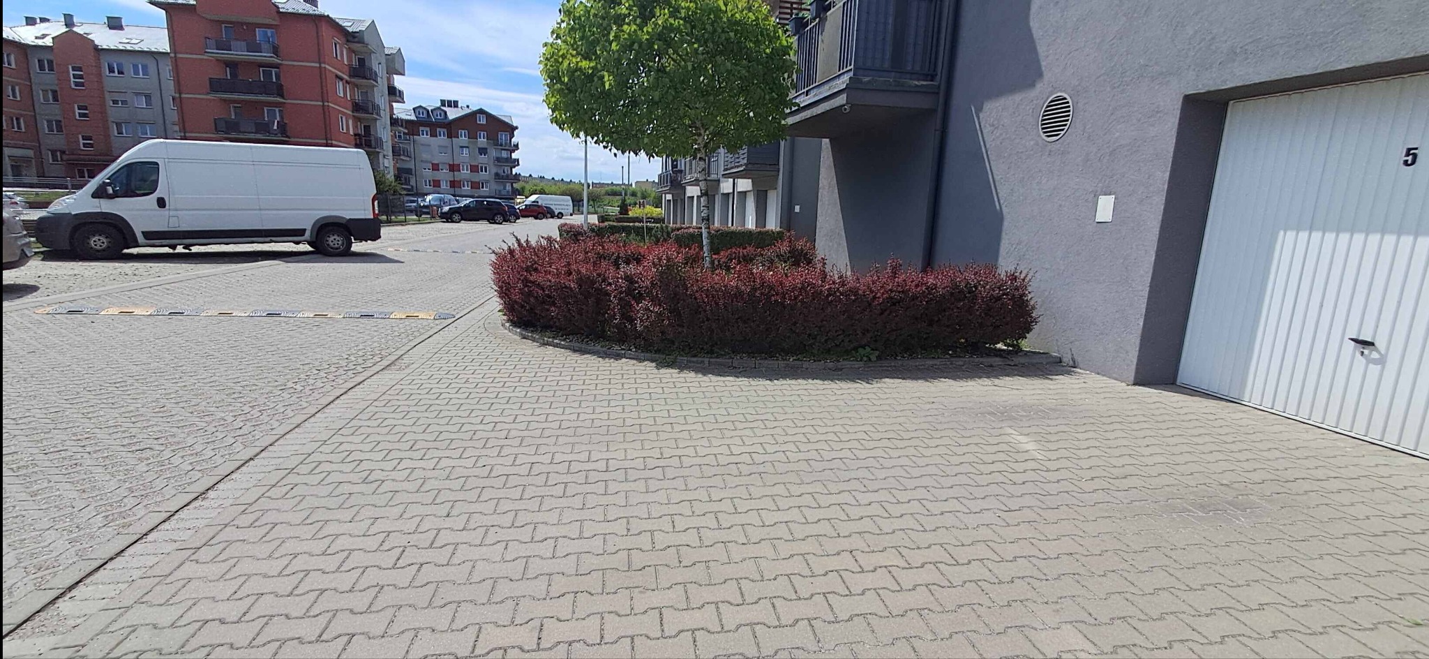 Kostka brukowa na podjeździe do garażu w nowoczesnym osiedlu, z widocznym białym vanem i starannie przyciętymi krzewami.