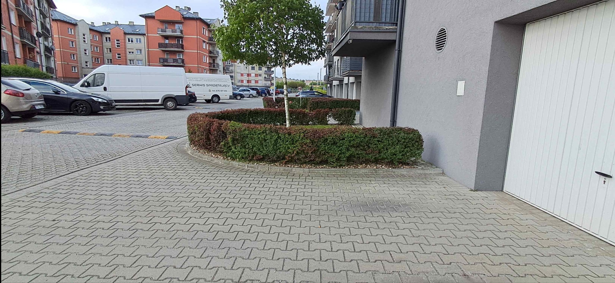 Szara kostka brukowa na parkingu osiedlowym z białym busem 'Serwis Sprzątający' w tle i zadbanym żywopłotem. Widok na budynki mieszkalne i garaże.