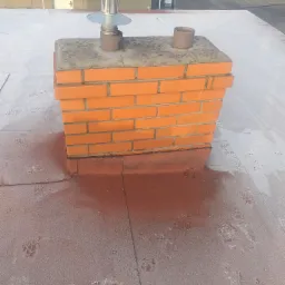 Murowany komin z metalowym zakończeniem na płaskim dachu pokrytym śladami butów i zabrudzeniami.