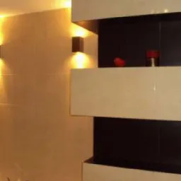 Nowoczesna łazienka z beżowymi płytkami i czarnymi akcentami. Oświetlenie punktowe podkreśla elegancki design. Widoczna wanna i dekoracyjne elementy na półce.