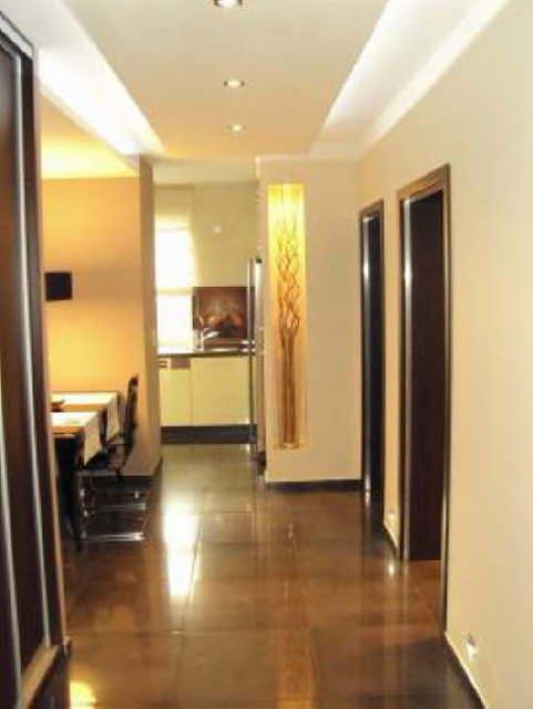 Wykończony korytarz z połyskującą podłogą, wnęką dekoracyjną i widokiem na kuchnię. Ściany w ciepłym, neutralnym odcieniu. Widoczne drzwi wewnętrzne.