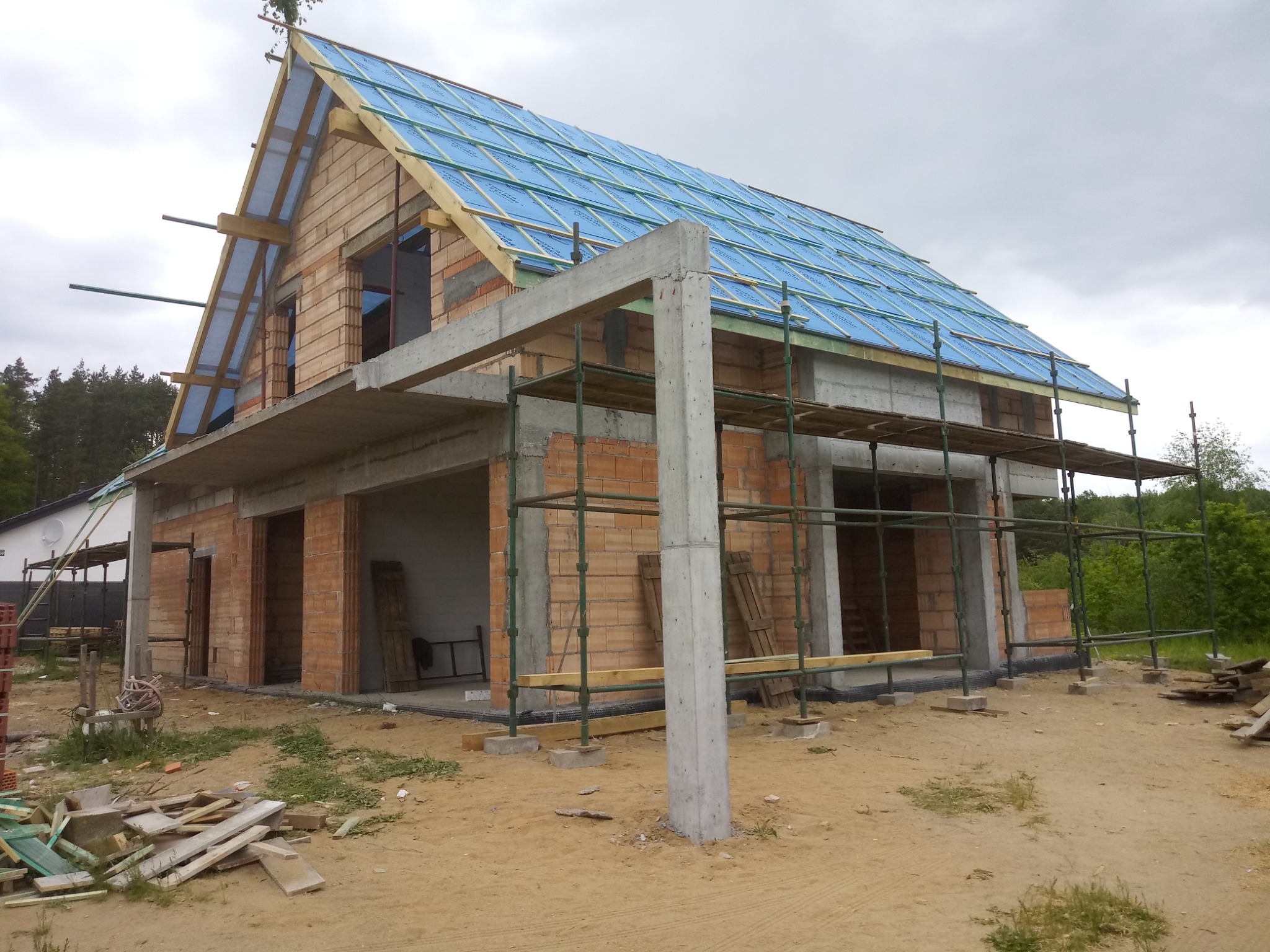 Surowy stan domu jednorodzinnego z cegły, z widocznym szkieletem dachu pokrytym niebieską membraną, otoczonego rusztowaniami i materiałami budowlanymi.