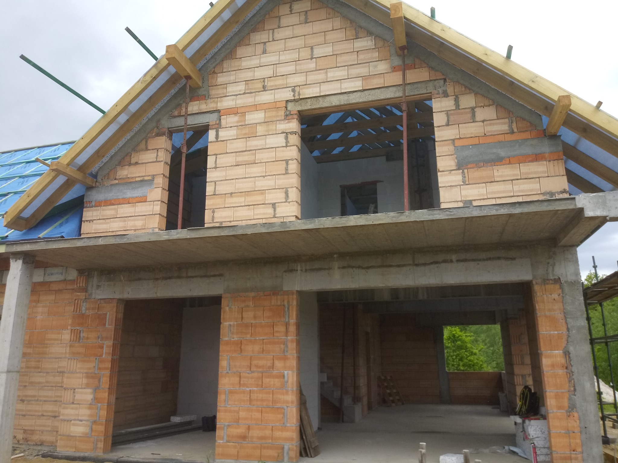 Surowy stan dwukondygnacyjnego domu z pustaków ceramicznych z widocznymi otworami okiennymi i zadaszeniem pokrytym folią budowlaną, betonowy strop między kondygnacjami, widoczne drewniane elementy...