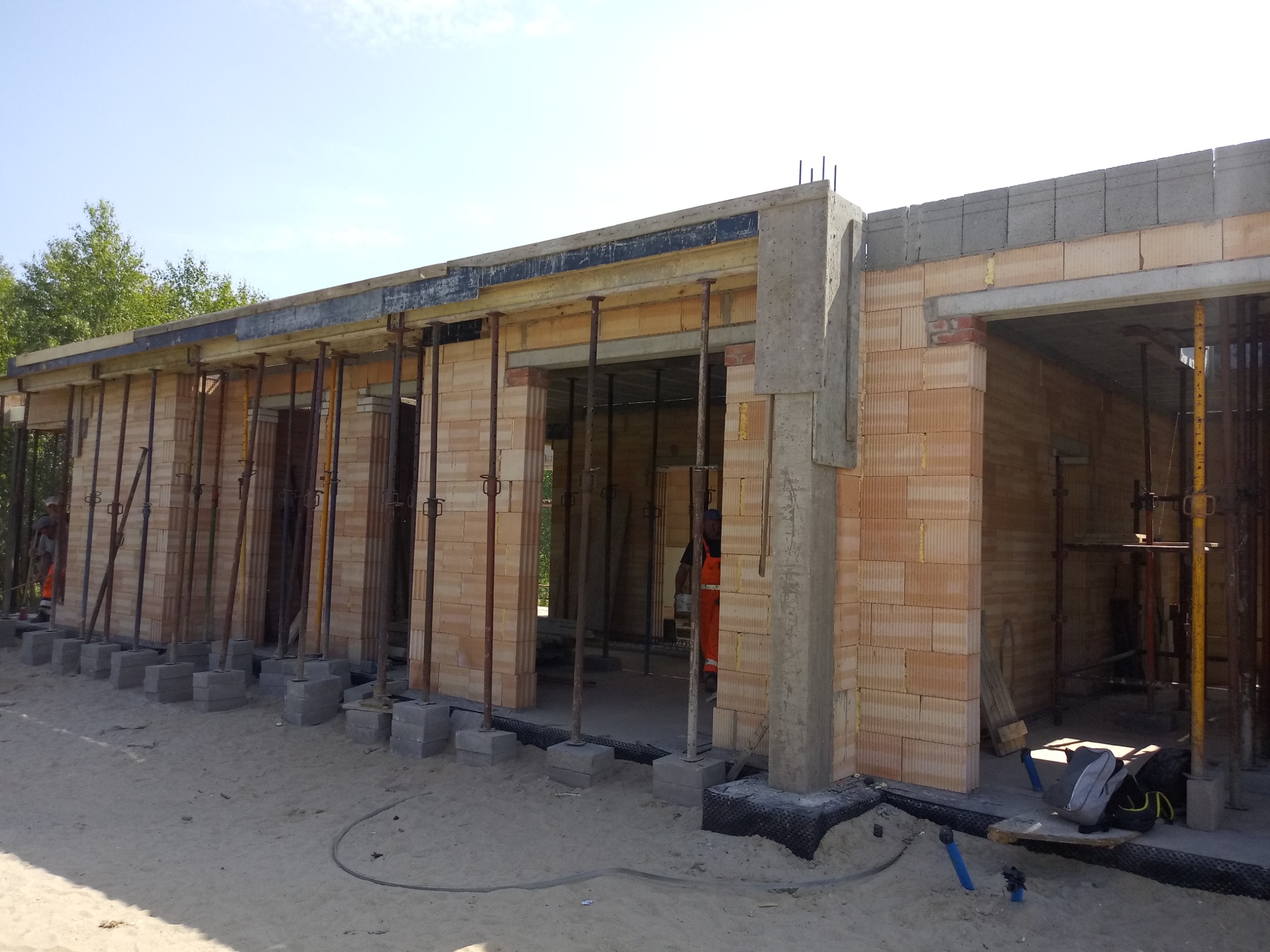 Surowy stan domu w trakcie budowy, widoczne ściany z pustaków ceramicznych, betonowe słupy, stalowe podpory, szalunki stropowe, teren budowy z piaskiem.