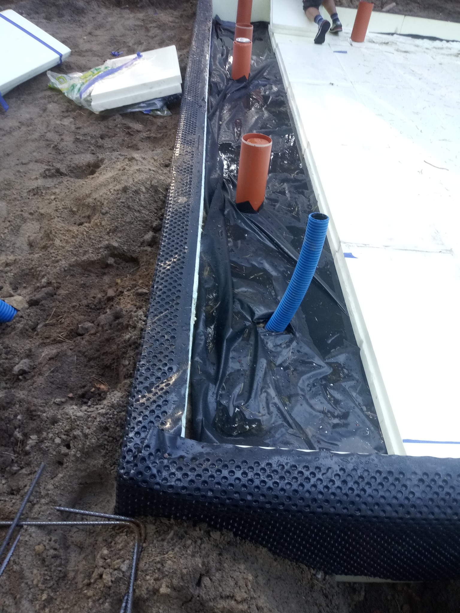 Przygotowanie fundamentów: izolacja pionowa z folii kubełkowej, rury kanalizacyjne w ziemi, białe płyty styropianowe, widoczne zbrojenie.