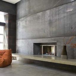 Perito development - Minimalistyczny salon z betonową ścianą, kominkiem i pomarańczowym fotelem. Widoczne duże okno z naturalnym światłem. Wazon z gałązkami jako element dekoracyjny.