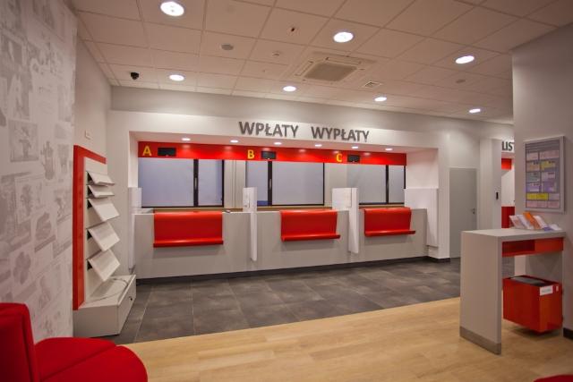 Jasne wnętrze placówki z ladami do obsługi klientów, oznaczone literami A, B, C, z napisem 'Wpłaty Wypłaty' powyżej. Czerwone akcenty, nowoczesny design.