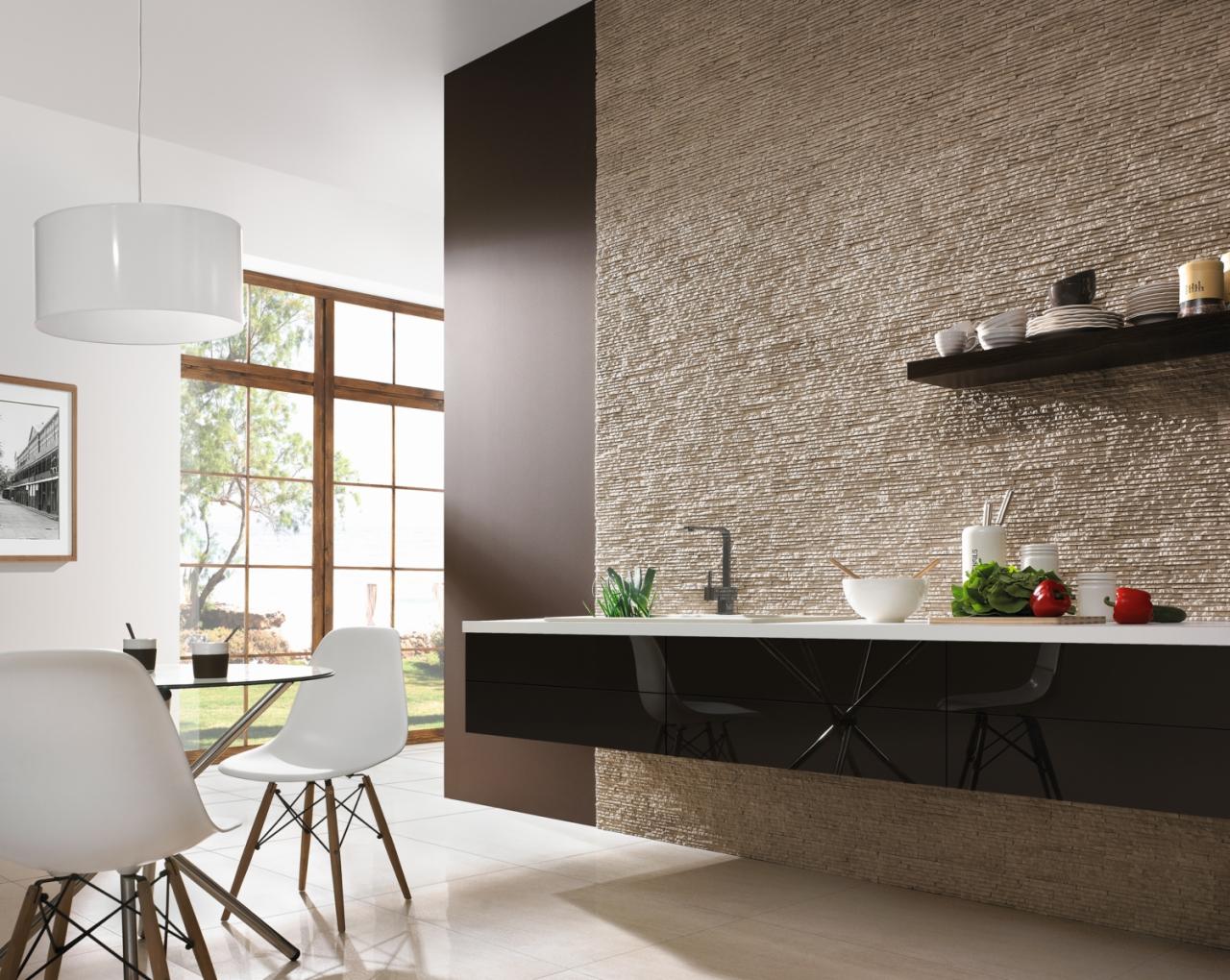 Nowoczesna kuchnia z dekoracyjną ścianą z kamienia, czarnymi szafkami i białym blatem. Minimalistyczne krzesła i okno z widokiem na zieleń dopełniają wystrój.