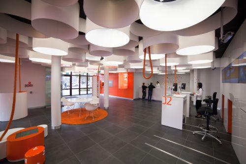 Nowoczesne wnętrze biurowe z designerskim oświetleniem sufitowym, pomarańczowymi akcentami i minimalistycznym wyposażeniem. Widoczni ludzie przy stanowiskach obsługi.