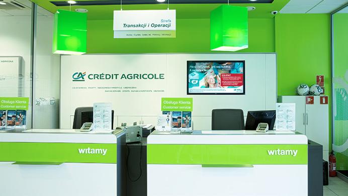 Wnętrze placówki bankowej Crédit Agricole z zielonymi akcentami: stanowiska obsługi klienta z napisem 'witamy', wiszące lampy i tabliczki informacyjne.