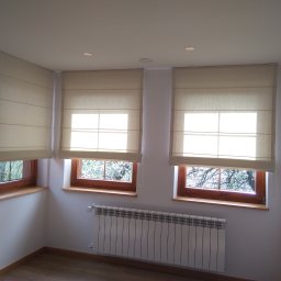 Twoje Okno Aranżacja i Dekoracja Okna Danuta Brzuchacz - Jasne, beżowe rolety rzymskie zamontowane w trzech oknach z drewnianymi ramami, widok na drzewa za oknem, białe ściany i grzejnik pod oknem.