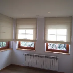 Jasne, beżowe rolety rzymskie zamontowane w trzech oknach z drewnianymi ramami, widok na drzewa za oknem, białe ściany i grzejnik pod oknem.