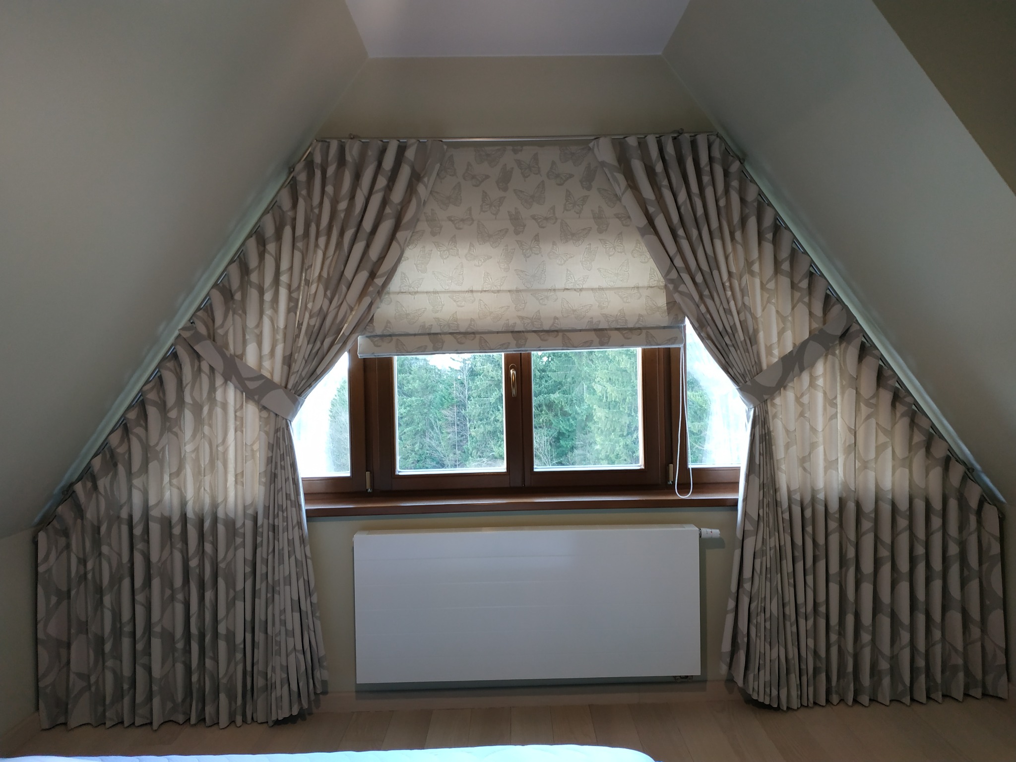 Zasłony w odcieniach szarości i beżu, upięte taśmami, z motywem motyli na roletach rzymskich w oknie na poddaszu.