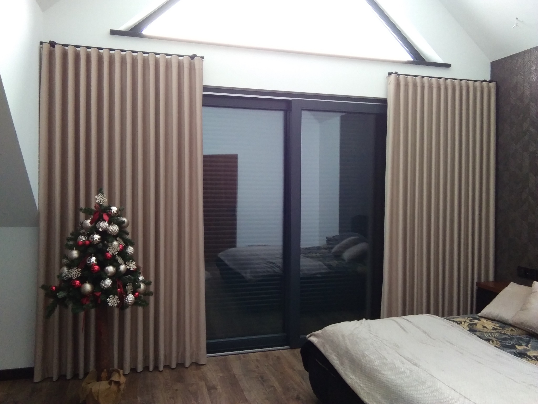 Beżowe zasłony w sypialni z oknem balkonowym i choinką w rogu, widok na łóżko przez szybę.