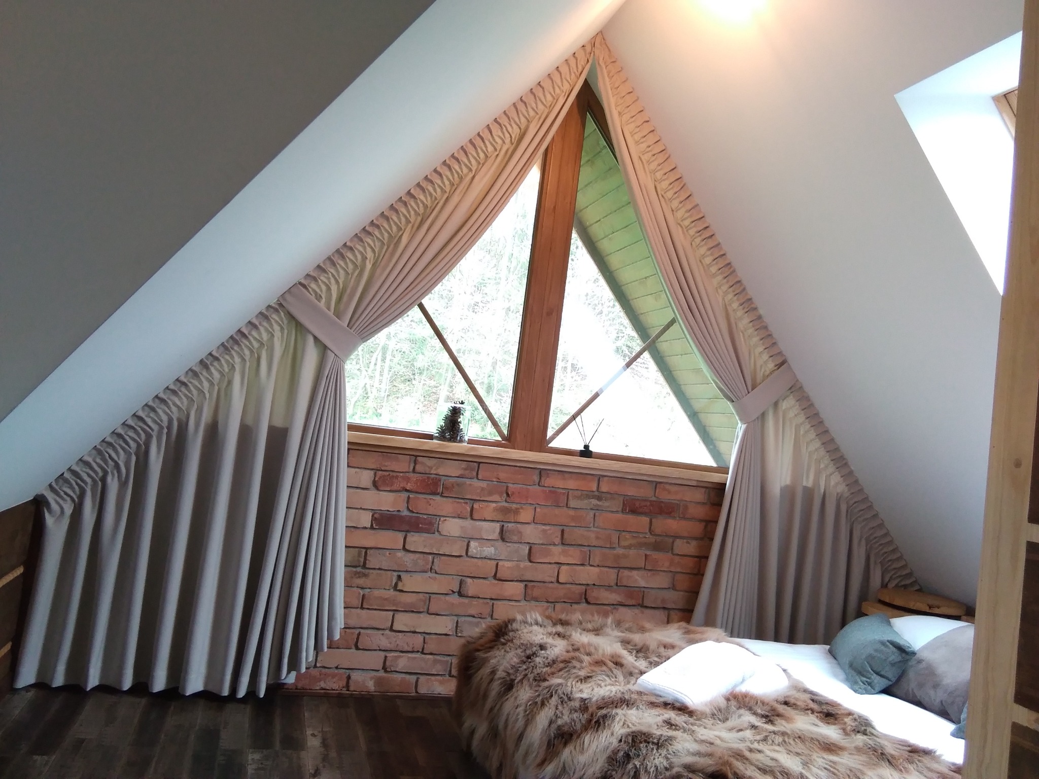 Beżowe zasłony w oknie o trójkątnym kształcie w pokoju na poddaszu z ceglaną ścianą i drewnianą podłogą.