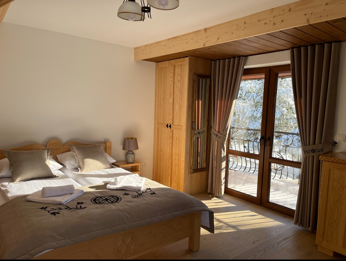 Jasny pokój w stylu góralskim z drewnianymi meblami, zasłony w kolorze beżowym upięte ozdobnymi pasami, widok na zaśnieżony krajobraz za oknem balkonowym.