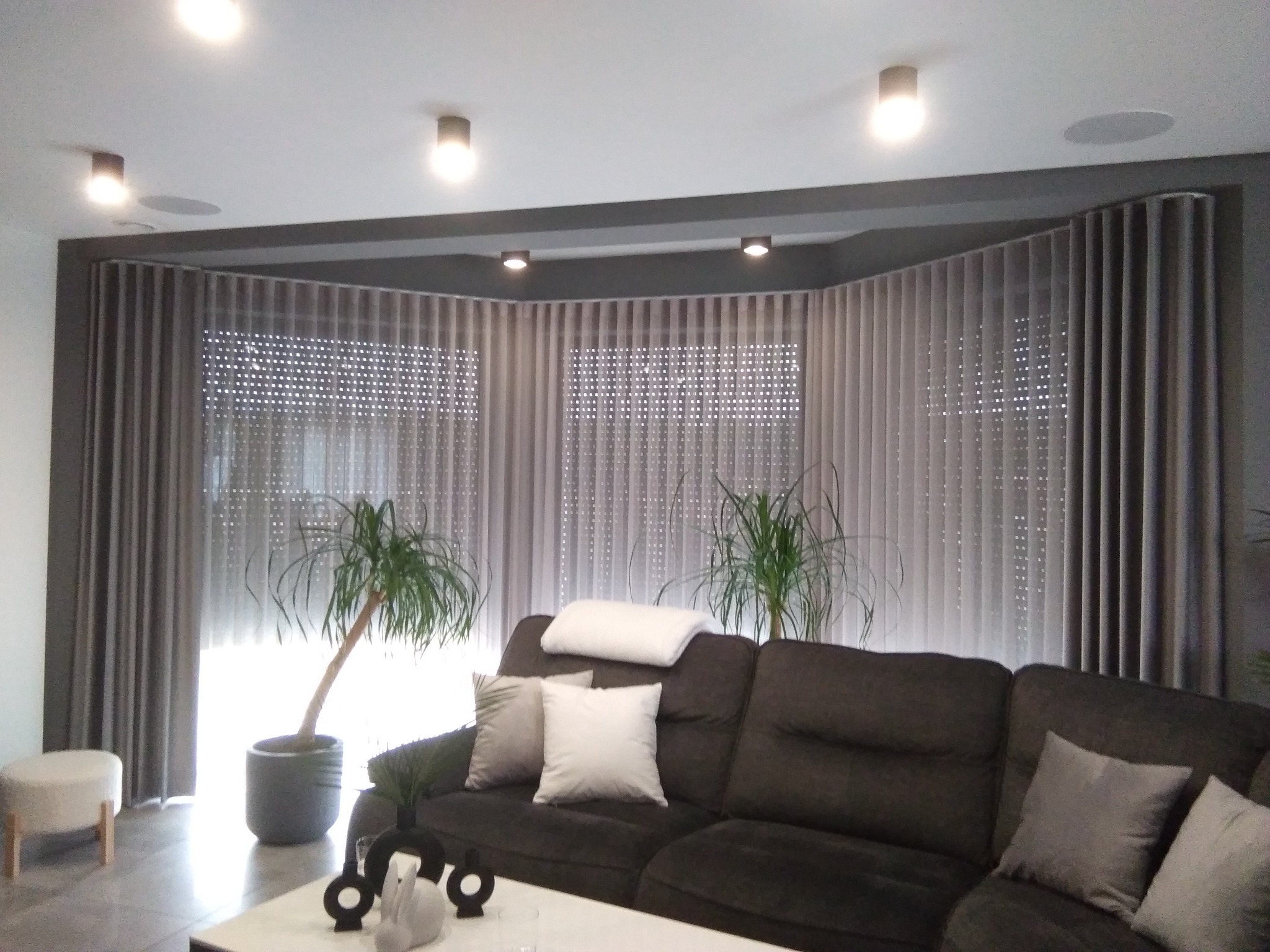 Elegancki salon z dużym oknem i szarymi, falującymi zasłonami na szynach, kanapa z poduszkami, rośliny doniczkowe i designerskie dekoracje na stoliku kawowym.