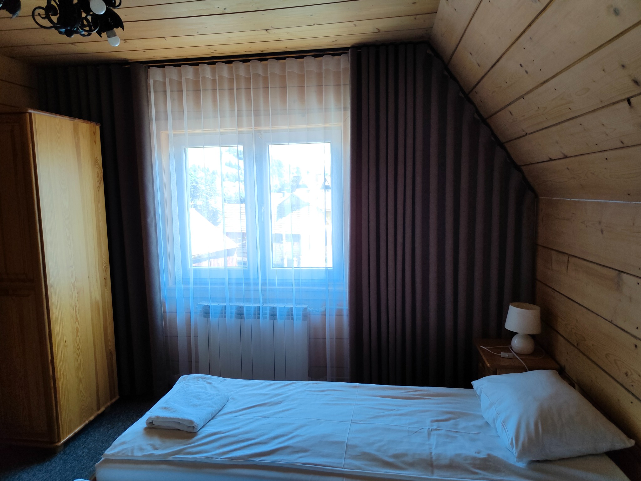 Pokój w drewnianym domku z zasłoniętymi oknami, brązowe grube zasłony po bokach okna i cienka, biała firanka na środku, widoczny grzejnik pod oknem.