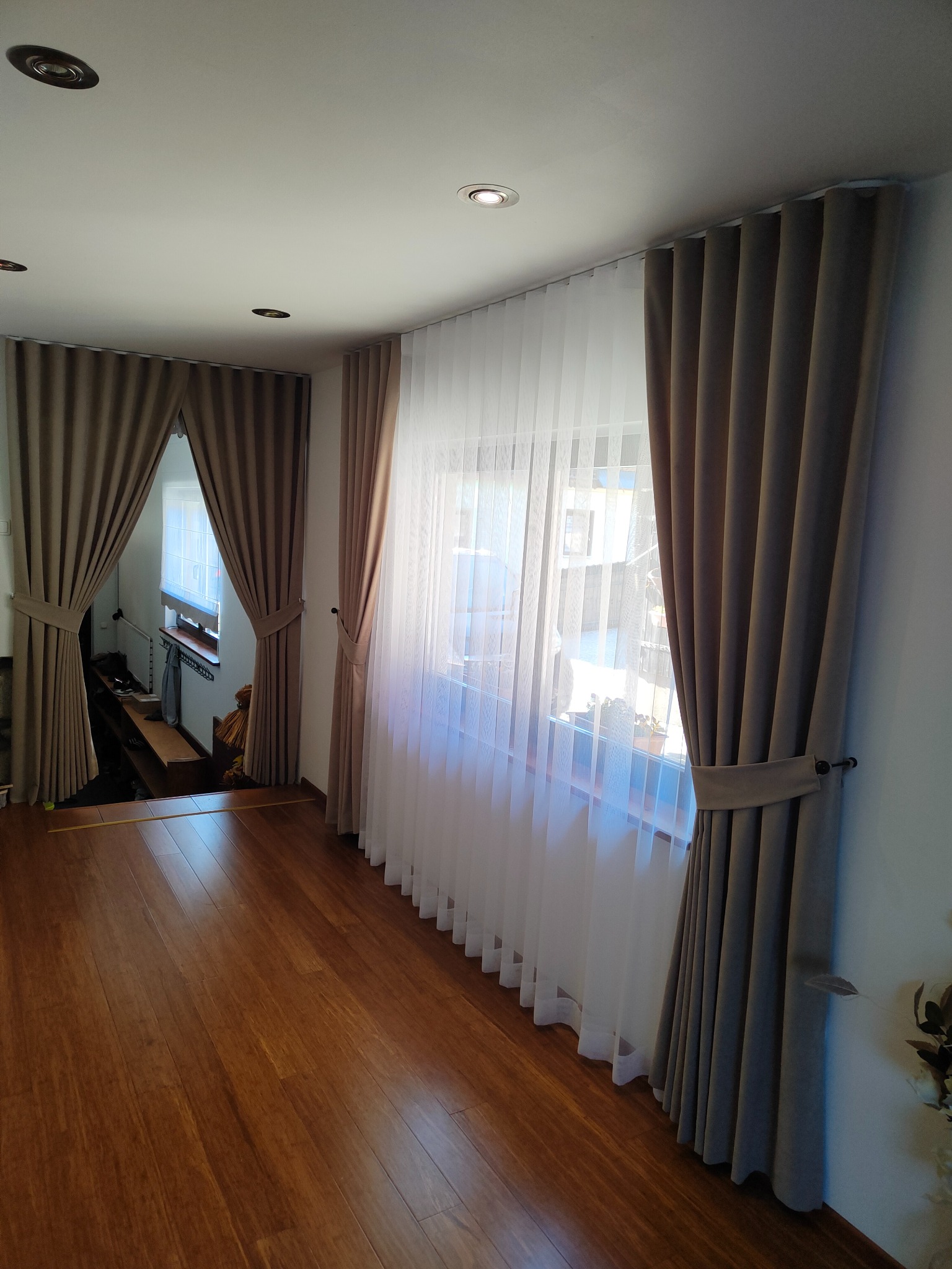 Eleganckie, długie, brązowe zasłony z podwiązkami i delikatne białe firany w przestronnym salonie z drewnianą podłogą, widoczne okno z roletą.