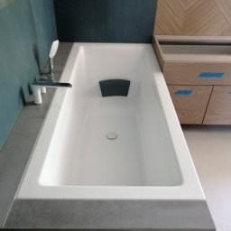 BANAŚ CONSTRUCTION Radosław Banaś - Nowoczesna, biała wanna z chromowaną armaturą, obłożona szarą płytką imitującą beton, z drewnianą zabudową w tle, zabezpieczoną niebieską taśmą malarską.