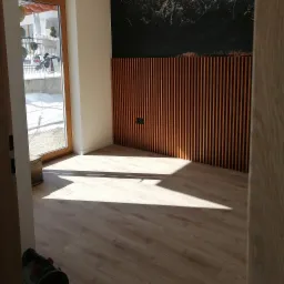 Jasne panele podłogowe w nowym pomieszczeniu, z dekoracyjną ścianą z drewnianych listew i czarnym panelem u góry, widok z perspektywy wejścia, wpadające światło słoneczne.