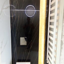 BANAŚ CONSTRUCTION Radosław Banaś - Część ściany w łazience wykończona czarnymi płytkami z złotymi żyłami, podświetlany okrągły przycisk spłuczki, fragment grzejnika drabinkowego i złota mozaika.