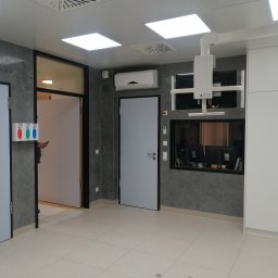 BANAŚ CONSTRUCTION Radosław Banaś - Wnętrze nowocześnie wykończonego pomieszczenia z szarą, teksturowaną ścianą, jasną podłogą z płytek, klimatyzatorem, drzwiami i sprzętem medycznym pod sufitem, widoczne monitory za szybą.