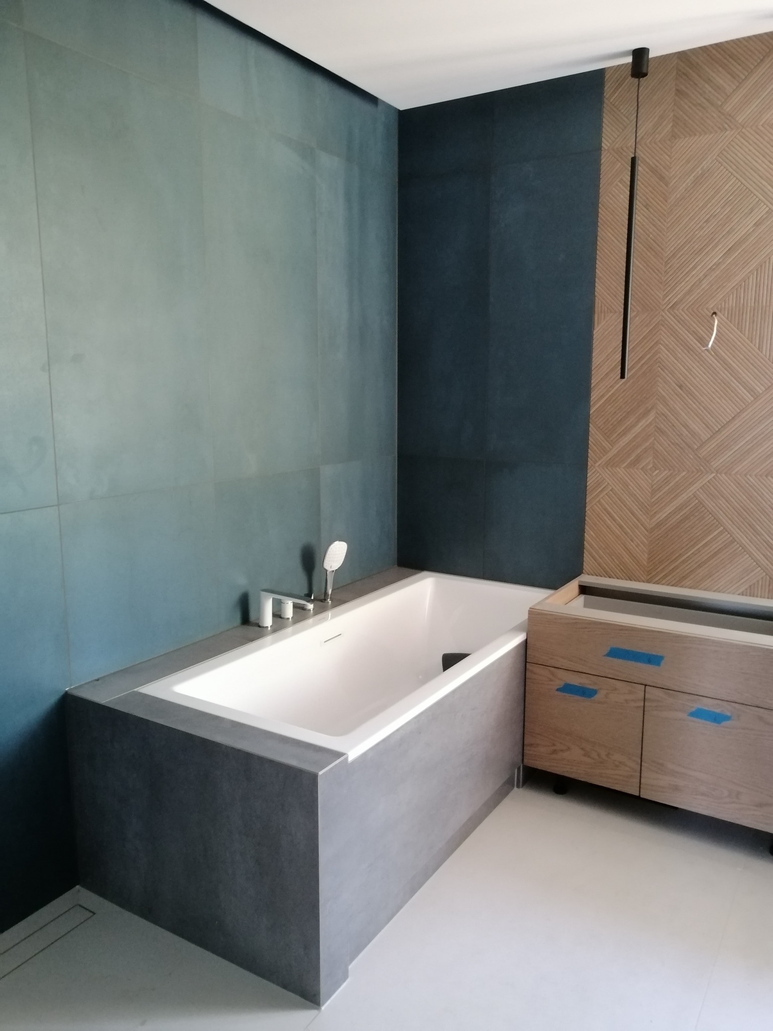 Nowoczesna łazienka z wanną obudowaną szarymi płytami, armaturą i szafką pod umywalkę z drewnianymi frontami zabezpieczonymi niebieską taśmą, ściany wyłożone dużymi, turkusowymi płytami...