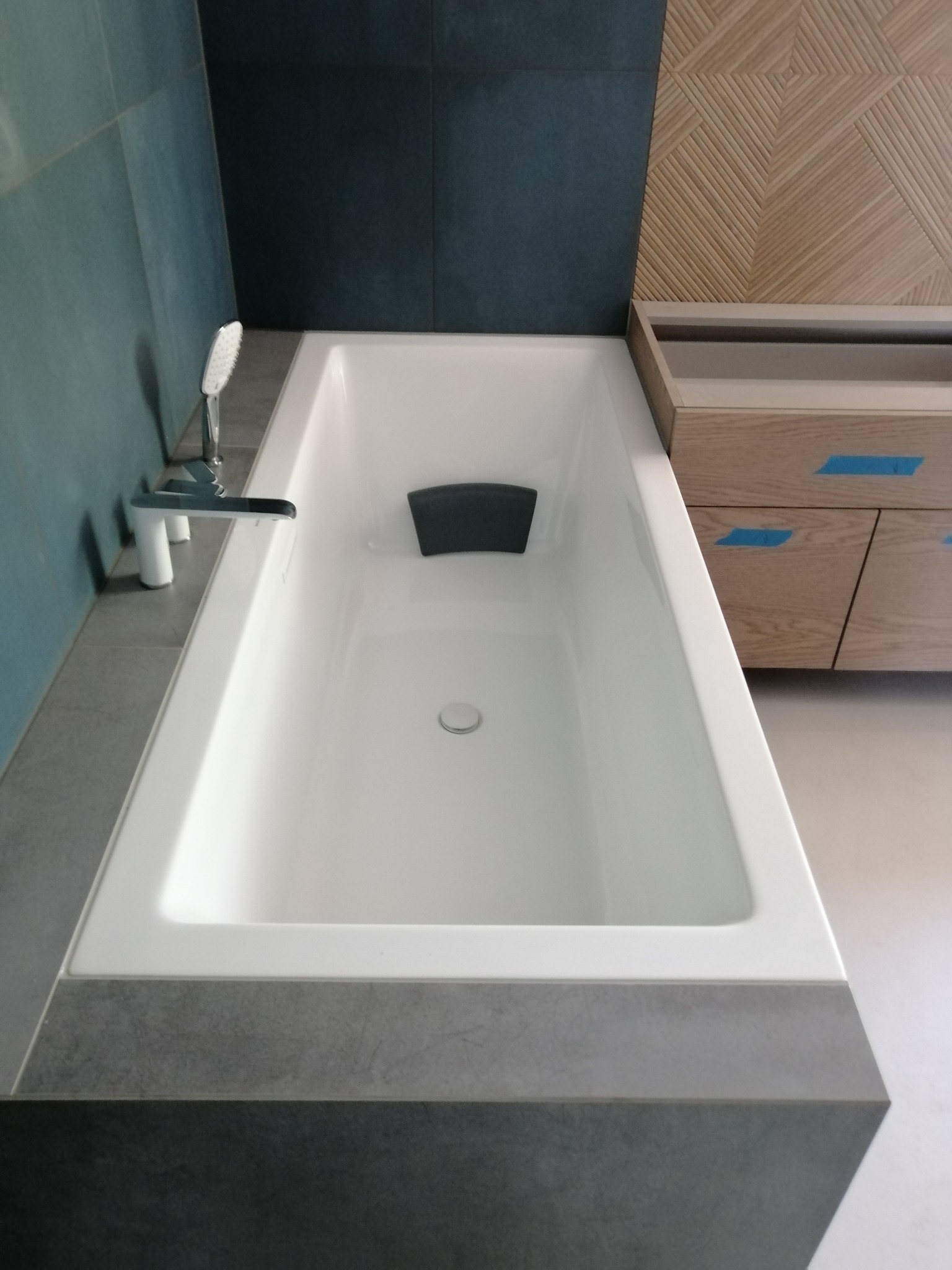 Nowoczesna, biała wanna z chromowaną armaturą, obłożona szarą płytką imitującą beton, z drewnianą zabudową w tle, zabezpieczoną niebieską taśmą malarską.