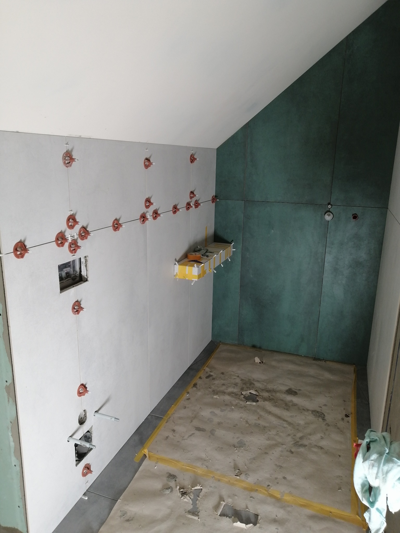 Wnętrze łazienki w trakcie prac wykończeniowych, widoczne białe płytki na ścianie z czerwonymi klipsami poziomującymi, fragment ściany pokryty zielonymi płytami, przygotowane podłoże pod posadzkę...