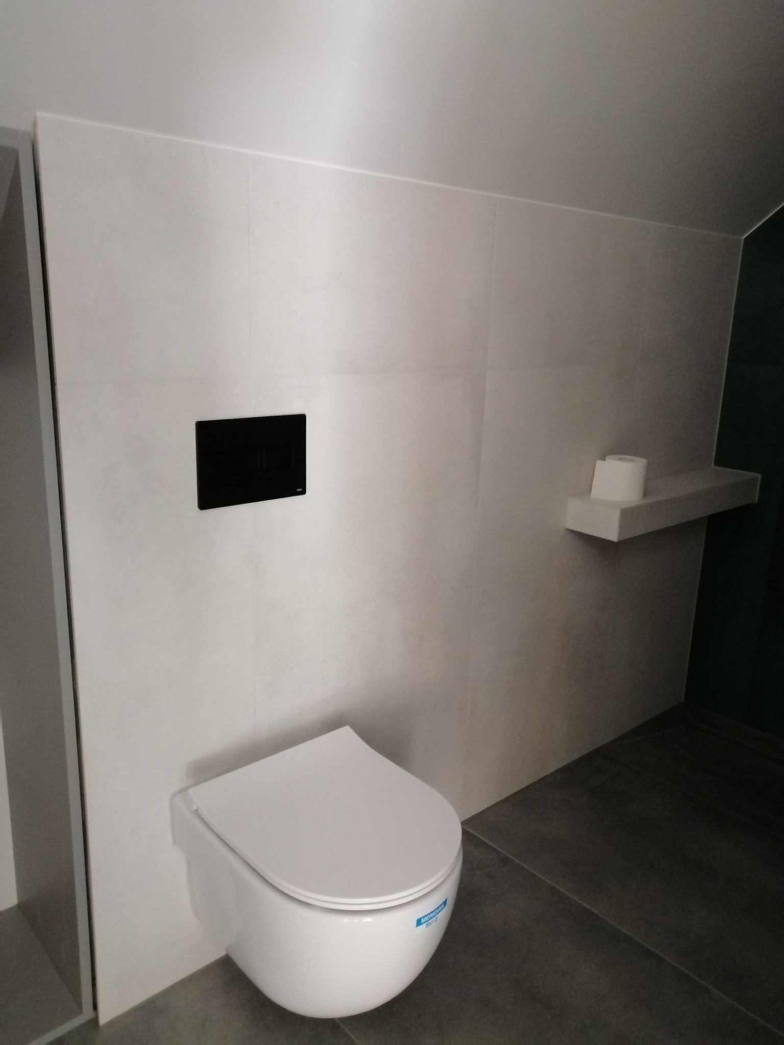 Nowoczesna, minimalistyczna łazienka z podwieszaną toaletą, szarymi płytkami i czarnym przyciskiem spłukującym na jasnej ścianie wyłożonej płytkami.