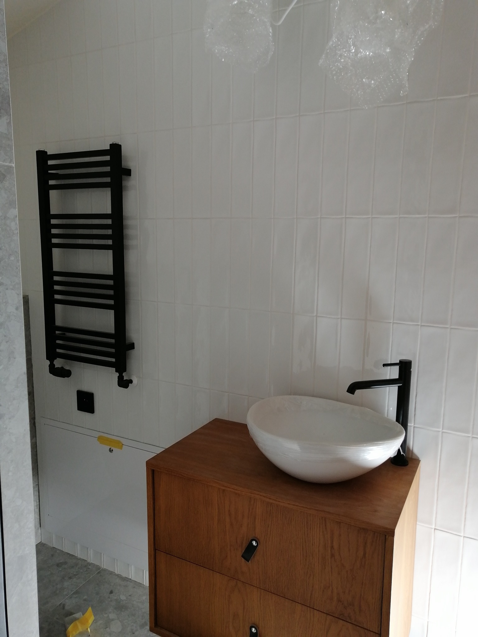 Nowoczesna łazienka z drewnianą szafką pod umywalkę, okrągłą białą umywalką na blacie, czarną baterią i grzejnikiem drabinkowym na białych, podłużnych płytkach; lampy wiszące zabezpieczone folią.