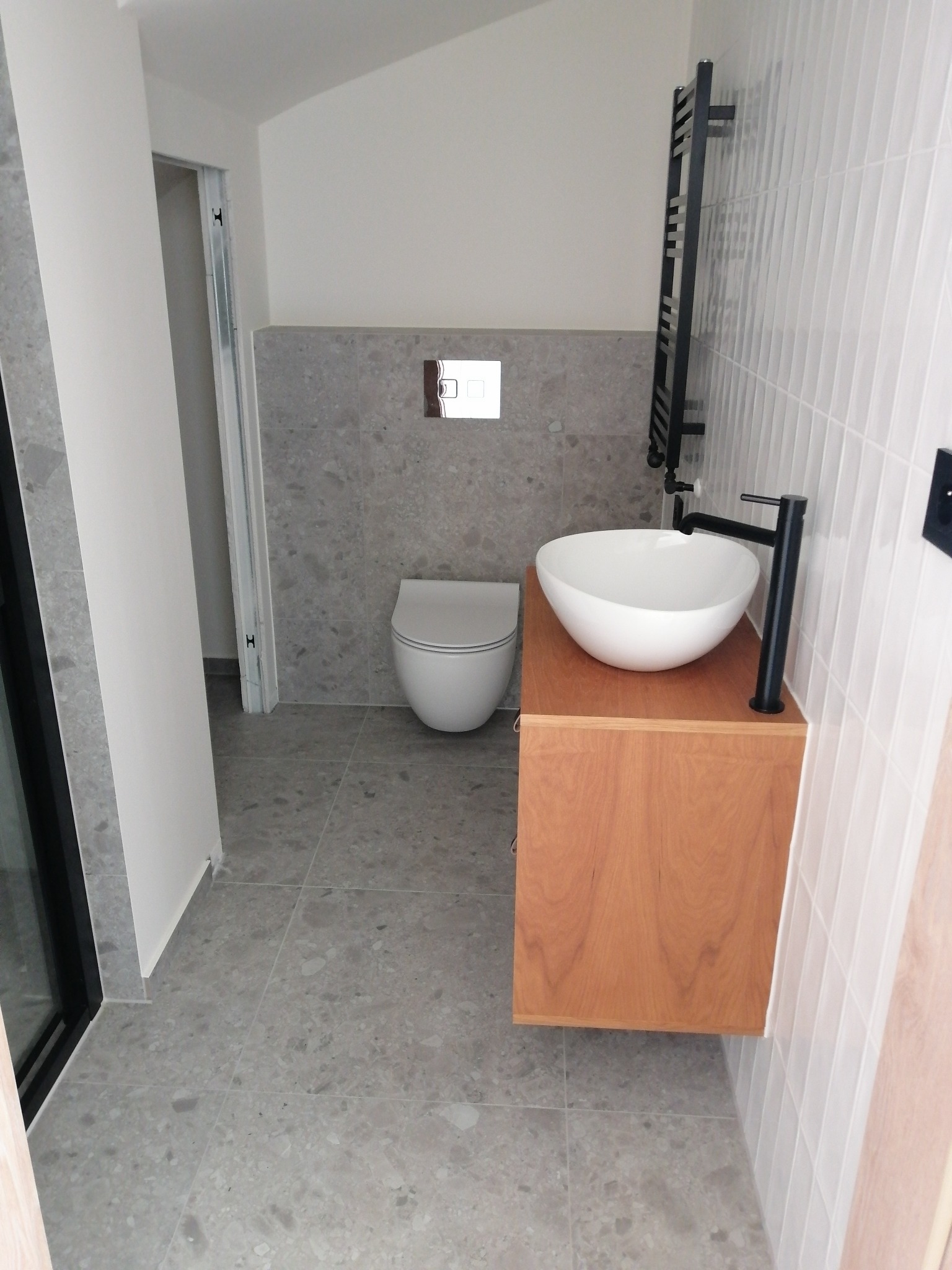 Nowoczesna łazienka z umywalką nablatową na drewnianej szafce, szarą mozaiką na ścianie z toaletą w tle i czarnym grzejnikiem drabinkowym.